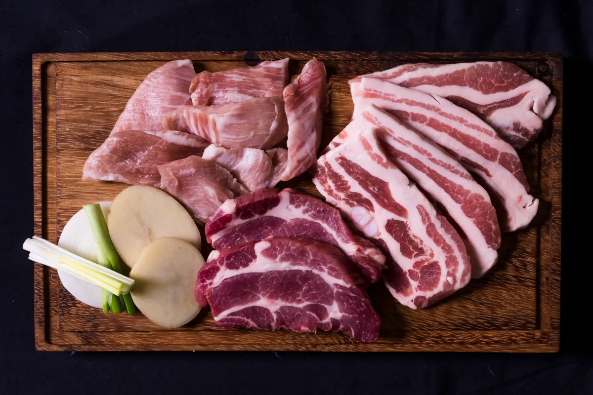 木のまな板にのった生の豚肉の数種類の部位と、玉ねぎ、ねぎ